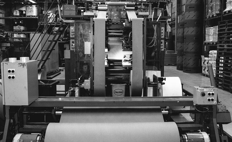 Industrial paper bag production machine in a factory, with large paper rolls and assembly components visible.